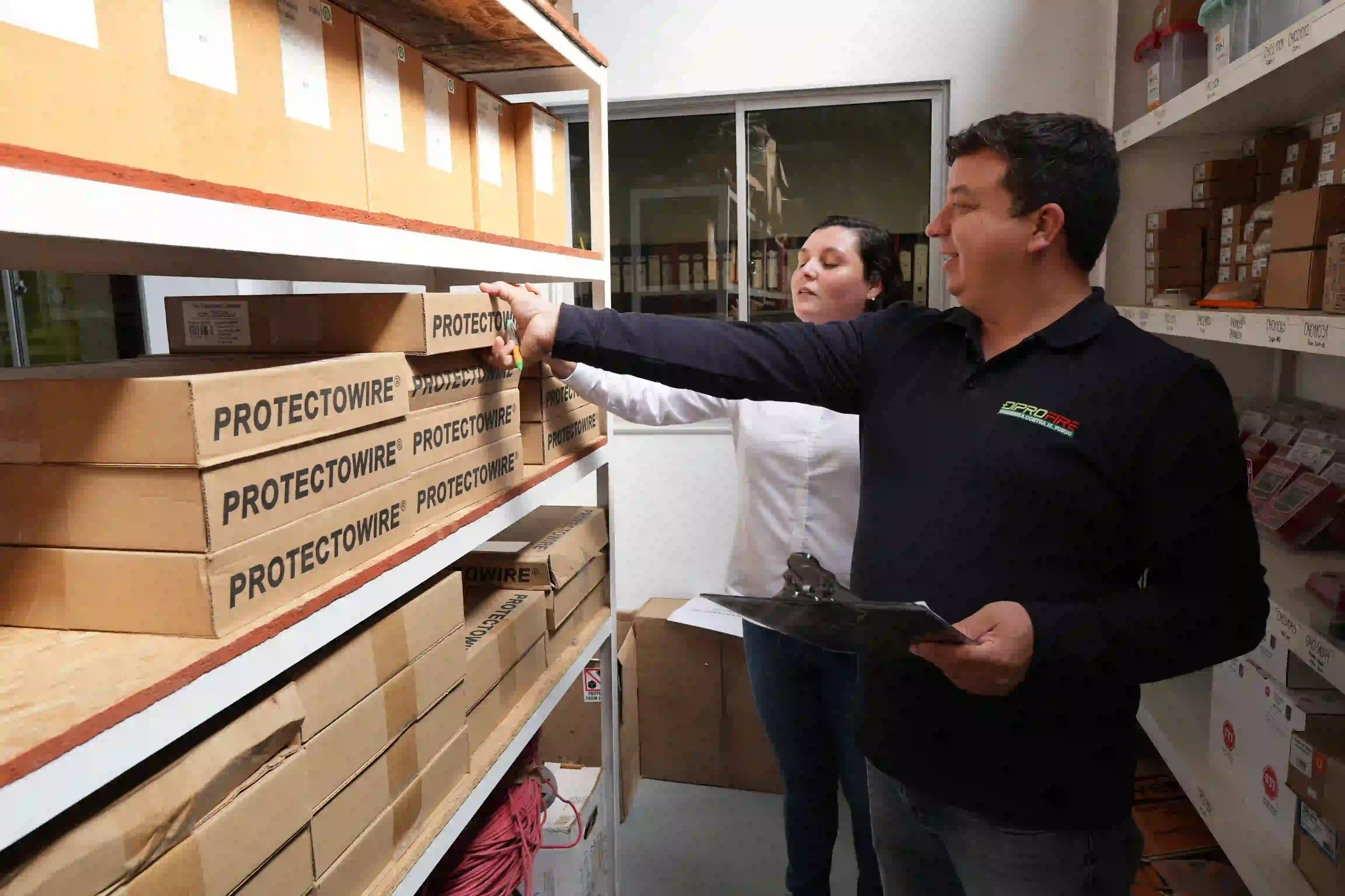 Dos miembros del equipo Diprofire organizando cajas de Protectowire en un almacén. Uno de ellos está tomando una caja de la estantería mientras el otro sostiene una tabla con un portapapeles.